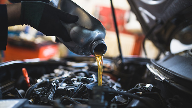 An individual in a workshop is pouring oil into an engine, with various car parts and tools visible around them.